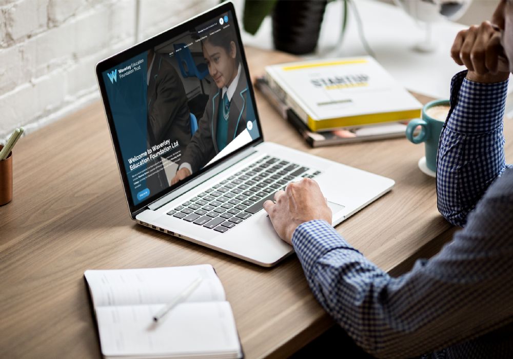 Waverley on a laptop on a desk, man with his hand to his chin, cup of coffee to the right. 